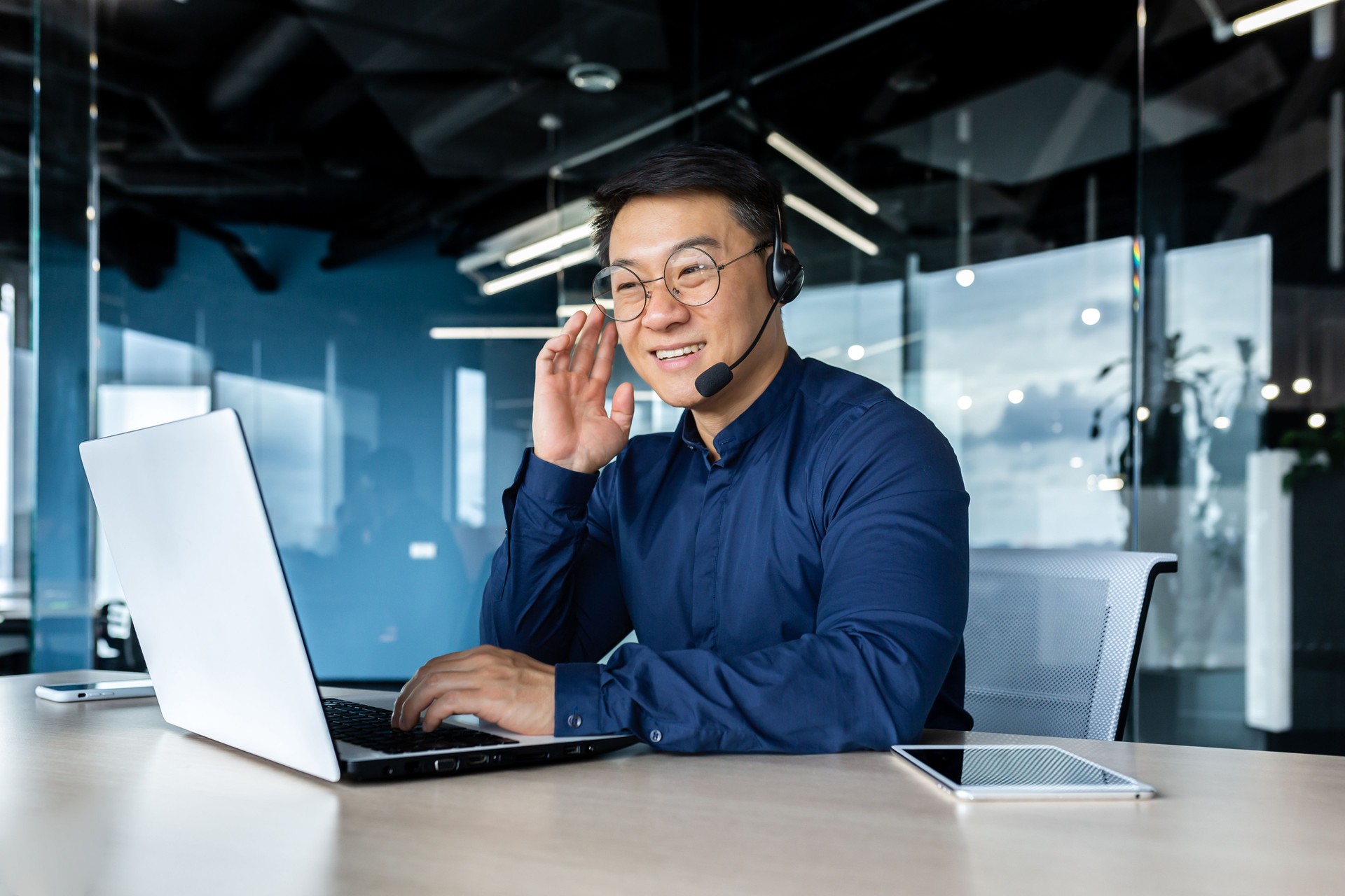 Cheerful and smiling office worker with video call headset, Asian customer support tech of online store, smiling and talking consulting. the man works inside the office